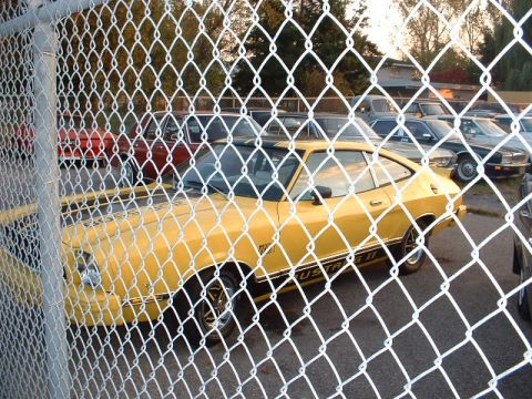 1978 Ford Mustang II - Yellow 1978 Ford Mustang II coupe seen through a chain-link fence. Other vehicles are parked nearby.