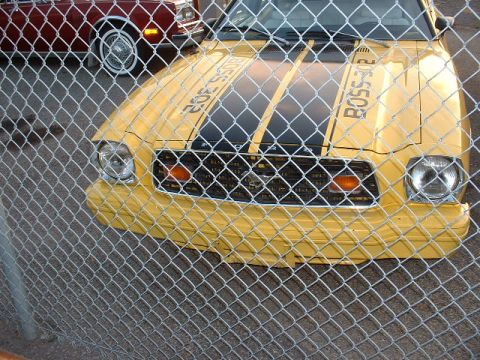 1978 Ford Mustang II - A yellow car with black racing stripes is parked behind a fence, with another vehicle partially visible in the background.