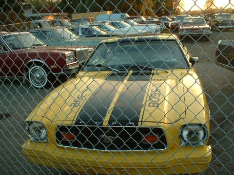 1978 Ford Mustang II - A yellow 1978 Ford Mustang II with racing stripes is parked behind a fence, with other vehicles visible in the background.