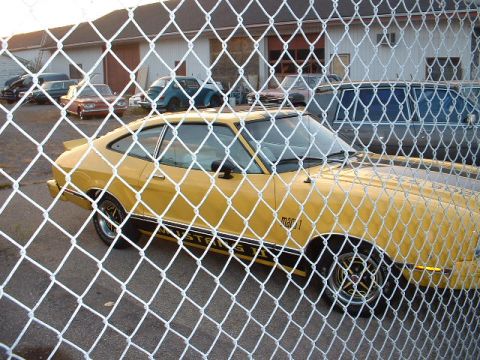 1978 Ford Mustang II - Yellow 1978 Ford Mustang II Mach 1 coupe parked behind a chain-link fence, with other vehicles visible in the background.