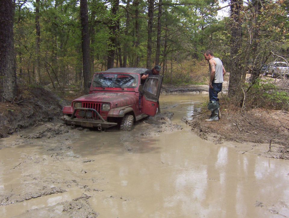 Red Jeep stuck in a muddy ditch with two guys around it. Another SUV is visible in the background