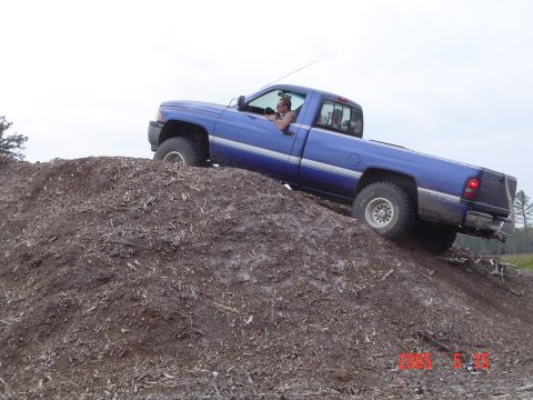 2005 Dodge 1500 - A blue pickup truck navigates an off-road obstacle, climbing a large pile of dirt.
