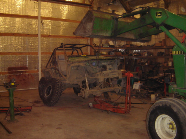 Off-road vehicle body with roll cage, missing engine and body panels, on jack stands in a workshop. A tractor loader is overhead