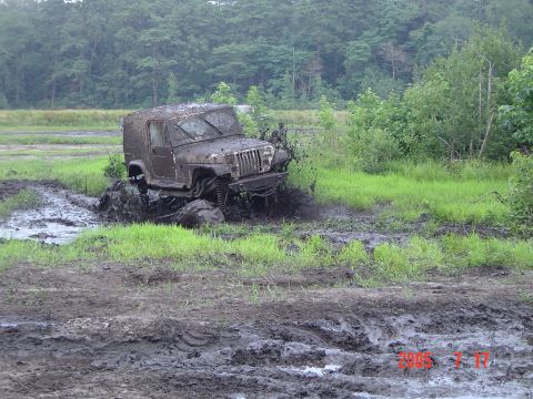 2005 Dodge 1500 - A Jeep Wrangler TJ navigates a challenging mud obstacle, kicking up a large spray of mud and water.