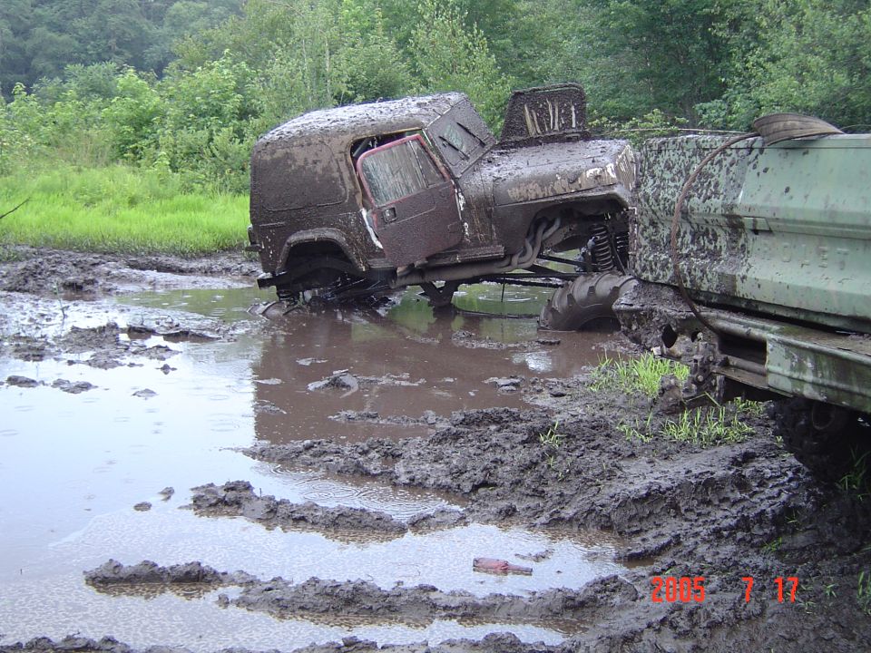 Two off-road vehicles stuck deep in mud and water, one is a brown Jeep-like vehicle with its door open, the other is a green truck