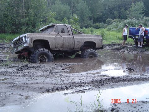 2005 Dodge 1500 - This heavily modified pickup truck is deeply stuck in a mud pit, with two onlookers standing by another vehicle in the background.