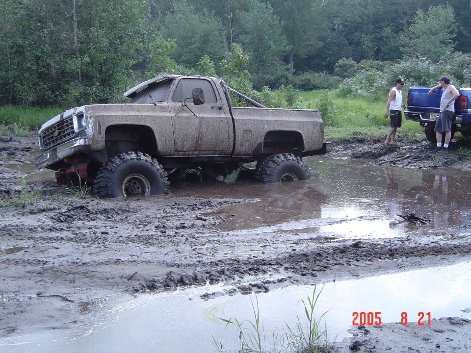 Mud-covered pickup truck stuck in a mud pit, with two people standing by a blue truck