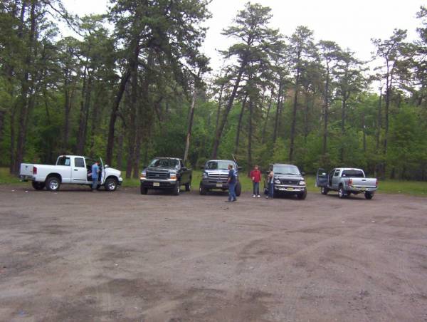 Several pickup trucks parked in a dirt lot with trees in the background