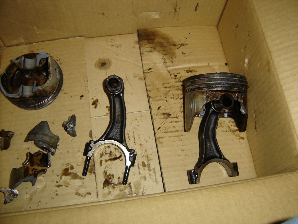 Damaged engine pistons and connecting rods in a cardboard box. One piston is broken, and pieces are scattered