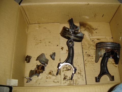 1988 Ford Bronco - Destroyed engine internals laid out in an oil-stained cardboard box: two pistons still attached to bent connecting rods, one with the crown…