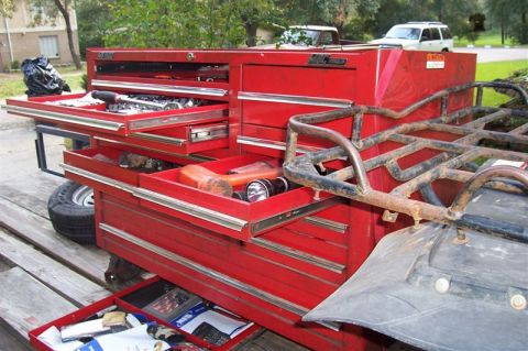 1987 Ford Bronco - A red toolbox filled with tools sits on a trailer, with a tire and ATV rack nearby. Two trucks are visible in the background.