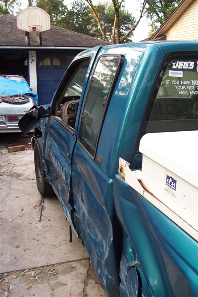 1987 Ford Bronco - A teal pickup truck with significant side damage is parked in a driveway next to another vehicle.