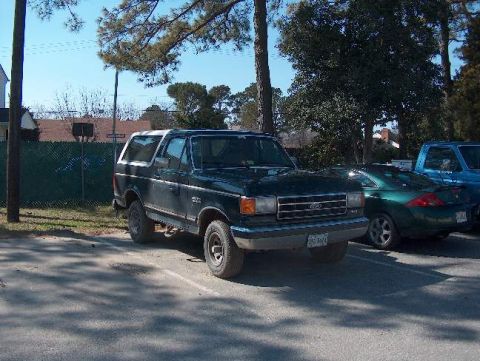 1991 Ford Bronco - A dark green Ford Bronco is parked in a lot with other vehicles under trees.