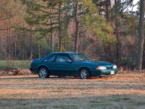 1993 Ford Mustang - A teal 1993 Ford Mustang is parked in a field with trees surrounding it.