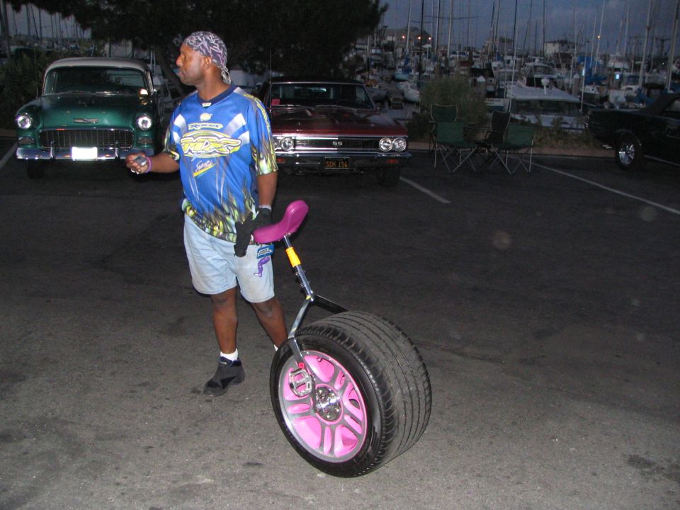 Man rolling a pink-rimmed tire with a bike seat past classic cars in a marina lot