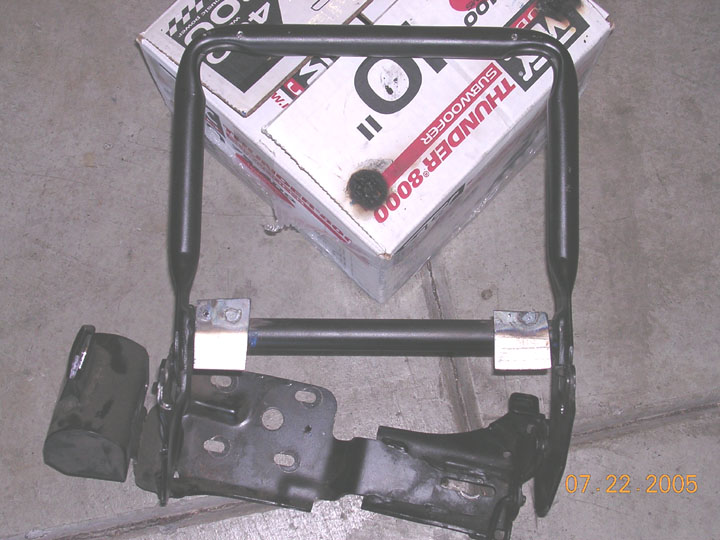 Black metal seat bracket assembly on concrete floor next to a subwoofer box