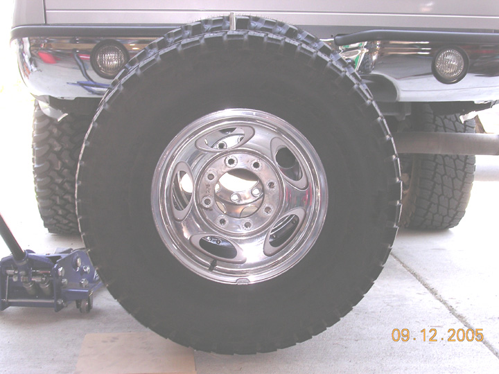Tall mud tire on polished 8-lug wheel standing beside a truck's rear bumper for size comparison