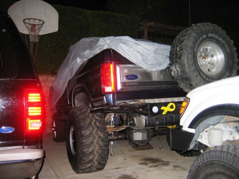 1985 Ford Bronco - A modified Ford Bronco with oversized tires is parked in a driveway at night, partially covered by a tarp.