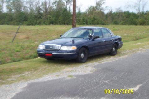 1998 Ford Crown Victoria - Dark blue Ford Crown Victoria sedan parked at an angle on a grassy shoulder next to a gravel drive, shot from the front three-quarter. It h…