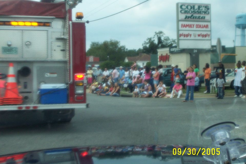 Fire truck parked on street with crowd watching a parade. Cole's Crossing Plaza sign in background
