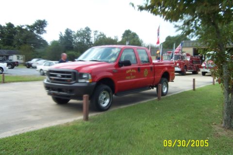 1998 Ford Crown Victoria - A red Ford F-Series pickup truck is parked next to other vehicles, including fire trucks, on a sunny day.