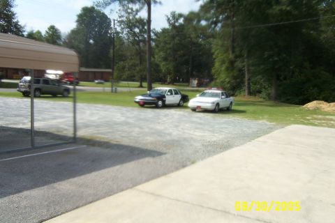 1998 Ford Crown Victoria - Two police vehicles are parked in a lot, one black and white and the other white, with trees and buildings in the background.