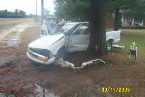 1998 Ford Crown Victoria - White pickup truck severely damaged after colliding with a tree on a wet, muddy road.