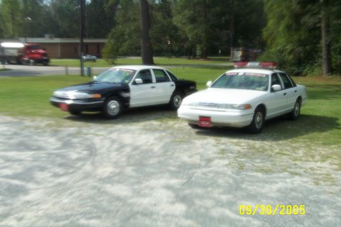 1998 Ford Crown Victoria - Two police cruisers, one white and one two-tone, are parked on a grassy area with trees in the background.