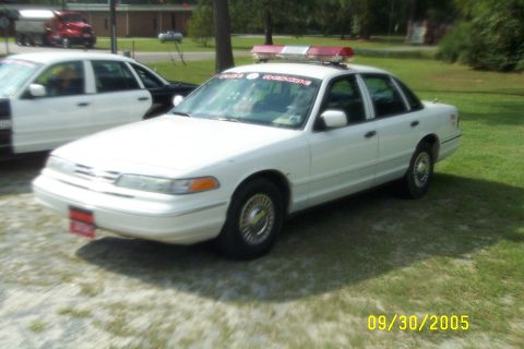 1998 Ford Crown Victoria - A white police car with a light bar is parked on grass next to another police car.