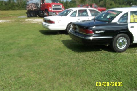 1998 Ford Crown Victoria - A black and white police car is parked on grass next to a white car, with a red truck in the background.