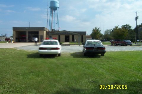 1998 Ford Crown Victoria - Two police cars are parked on the grass in front of a fire station building.