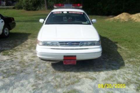 1998 Ford Crown Victoria - A white 1998 Ford Crown Victoria police car is parked outside, with another vehicle visible to the left.