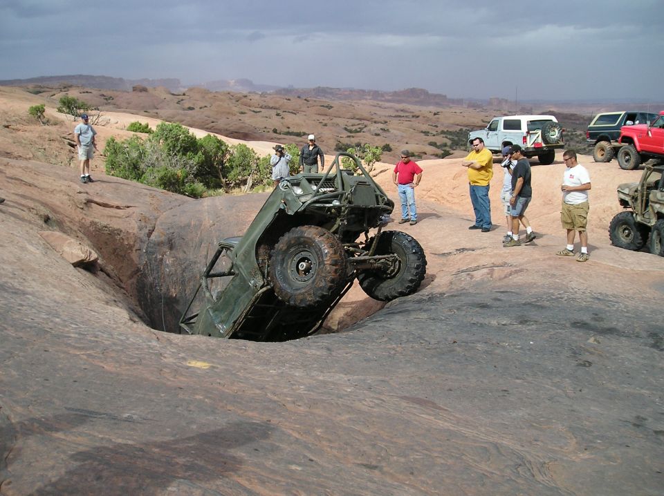 Green off-road vehicle tilted into a rock hole with people watching. Other vehicles are parked nearby
