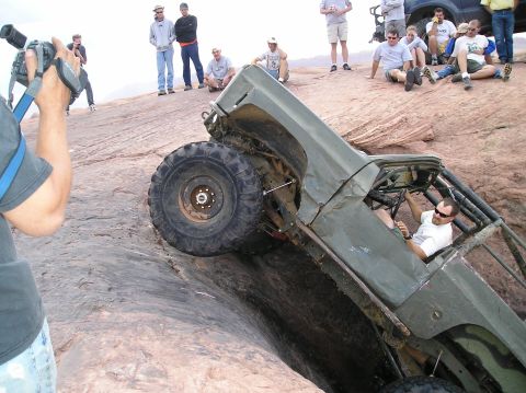 1993 Ford Bronco - A modified off-road vehicle is stuck in a deep crevice on a rocky terrain, with onlookers observing the situation.