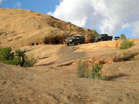 1993 Ford Bronco - Several SUVs navigate a challenging off-road path through a desert landscape under a partly cloudy sky.