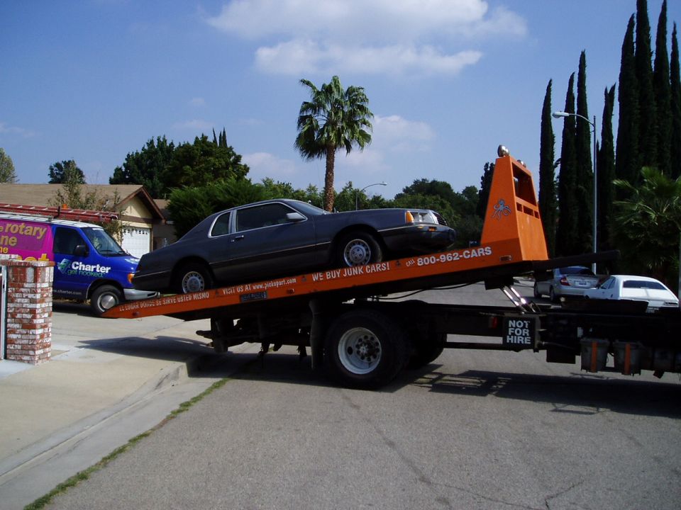 Gray Ford Thunderbird coupe loaded on an orange 'We Buy Junk Cars' flatbed tow truck on a sunny street