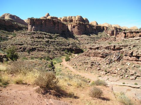 1993 Ford Bronco - A dirt road cuts through a desert landscape with rocky cliffs and sparse vegetation under a bright blue sky.
