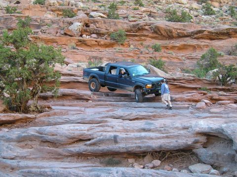 1993 Ford Bronco - A dark blue pickup truck navigates a challenging rocky trail in a desert landscape, with a person on foot providing guidance.