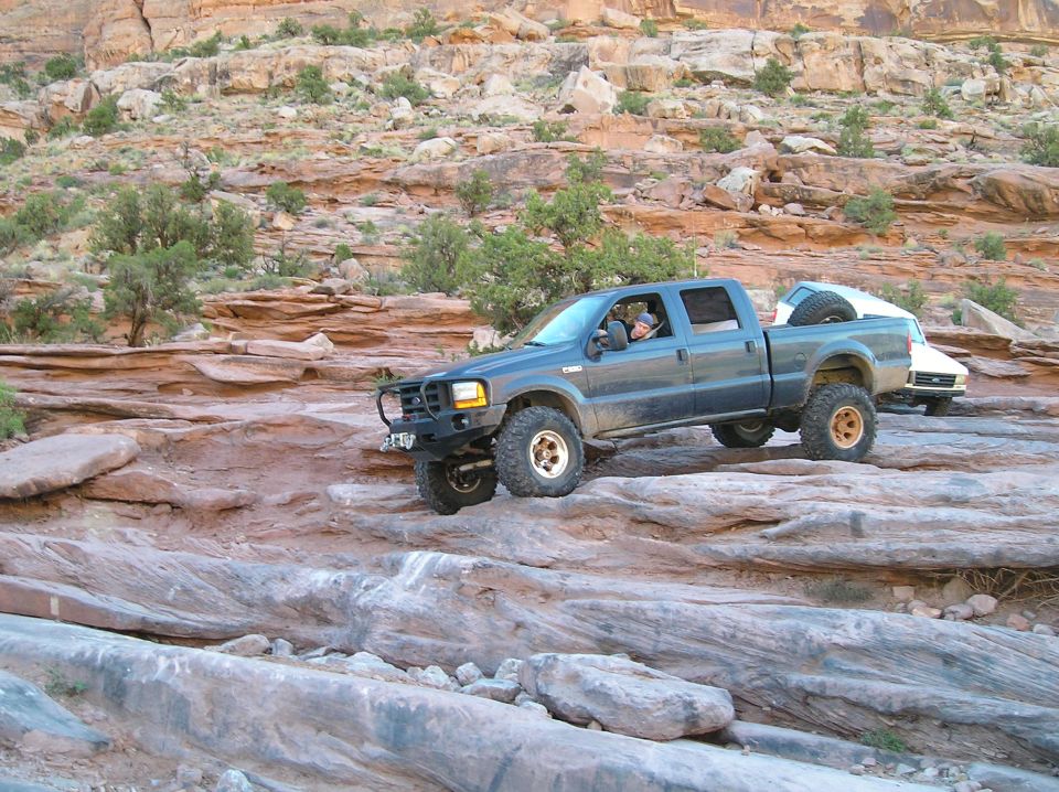 Dark blue Ford F-Series pickup truck with aftermarket bumper and spare tire on roof rack, driving over rocks. White vehicle behind it