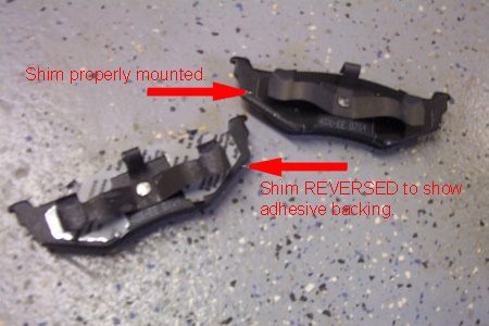 1999 Chrysler 300M - Two disk brake pads are shown, one with a properly mounted shim and the other reversed to reveal its adhesive backing.