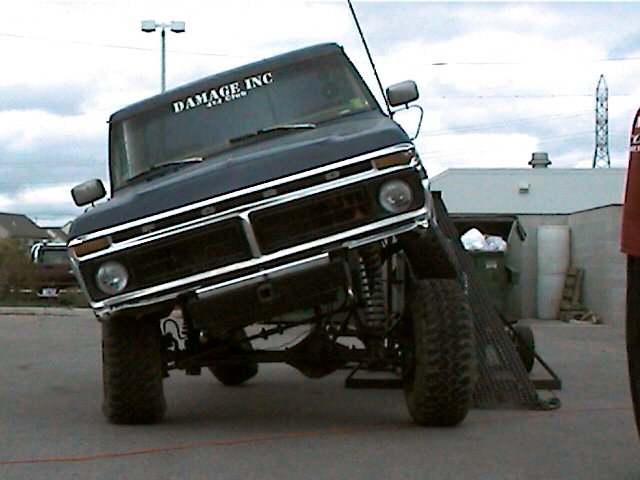Ford F-150 tilted on a ramp, showing its suspension and front grille. Sticker on windshield says 'DAMAGE INC'
