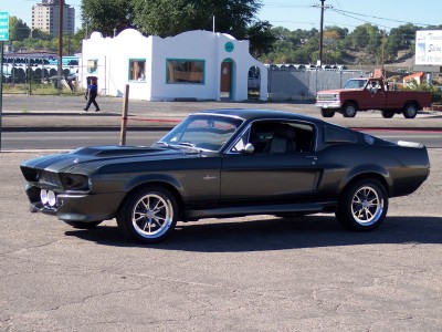1972 Ford Ranchero - Dark 1967 Ford Mustang Fastback with hood scoop and aftermarket wheels sits parked.