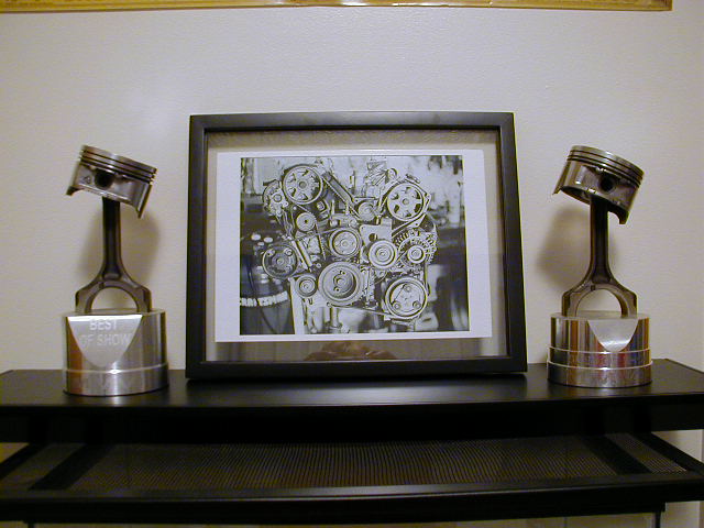 Framed photo of an engine's timing belt system, flanked by two piston trophies labeled 'Best of Show'