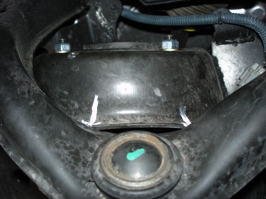 Underbody view of a suspension mount with two bolts, a green-marked bushing, and white alignment marks