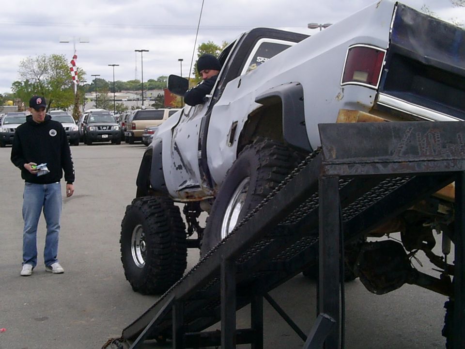 Gray lifted pickup truck with large tires on a metal ramp, driver visible, another person standing nearby