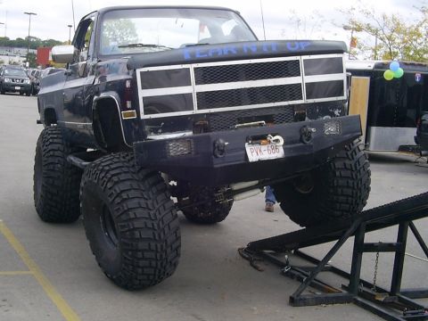 1985 Ford Bronco - A heavily modified pickup truck with oversized tires and a custom front bumper is parked outdoors, with a trailer in the background.