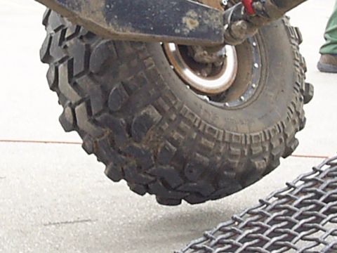 1985 Ford Bronco - Detail of an off-road tire and suspension system, with metal grating in the foreground.