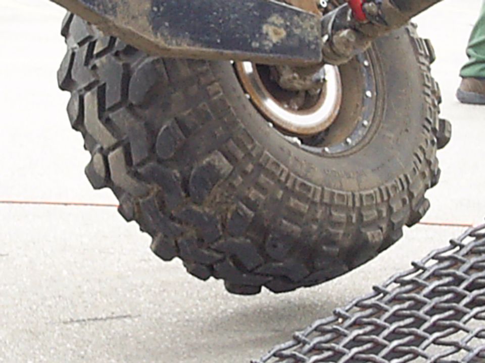 Close-up of a large off-road tire with aggressive tread, attached to a vehicle's suspension. Metal grating is visible in the foreground