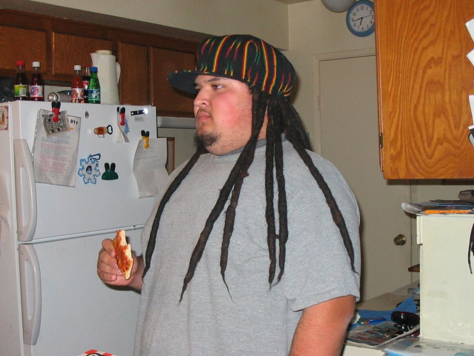 Person in a Rasta cap with dreadlocks holding a pizza slice in a home kitchen