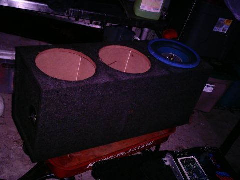 1986 Ford Bronco - A carpeted dual-12 subwoofer box sits on a red Radio Flyer wagon in a dim garage, its two bare MDF baffle rings and flush-mount terminal cu…